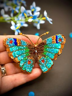 a hand holding a butterfly brooch in it's right hand and blue flowers behind it