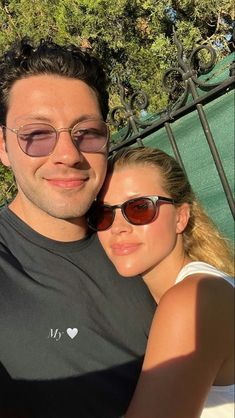 a man and woman taking a selfie in front of a green fence with trees behind them