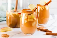 two glasses filled with apple cider on top of a white table next to cinnamon sticks