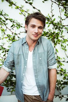 a young man standing in front of a wall covered with green leaves and branches, looking at the camera