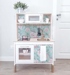 a white wooden play kitchen with floral wallpaper and potted plants on the shelves