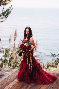 a woman in a red dress holding a bouquet on a wooden deck near the ocean