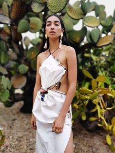 a woman in a white dress standing next to a cactus plant with large earrings on her head