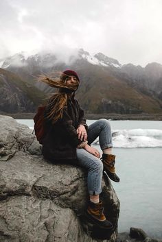 a woman sitting on top of a rock next to a body of water with mountains in the background