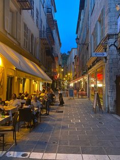 people are sitting at tables in an alleyway with buildings on either side and one person walking down the street