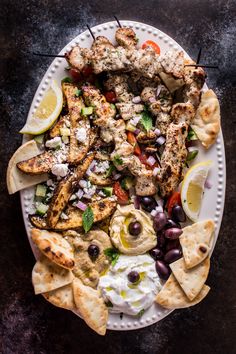 a white plate topped with chicken and pita bread next to olives, cucumber, and lemon wedges