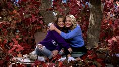 two women hugging each other in front of trees with red leaves on them and one woman is wearing a blue sweater