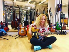 a woman is sitting on the floor with her guitar in front of her and many guitars behind her