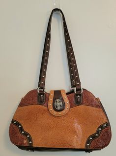 Tooled leather in shades of brown with silver hardware add the western flare. This bag has many pockets and pouches to help with organization. Silver studded double handle has a comfortable 12.5" drop. This sturdy and stylish bag is a great value. Brown Satchel Shoulder Bag With Silver-tone Hardware, Brown Double Handle Satchel With Snap Closure, Brown Shoulder Bag With Silver-tone Hardware For Everyday, Black Evening Bag, Carryall Tote, Guess Bags, Brown Leather Bag, Brown Purses, Cow Girl