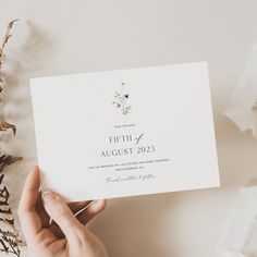 a person holding up a wedding card on top of a table with flowers and greenery