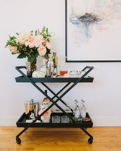 a bar cart with drinks and flowers on it in front of a wall mounted painting