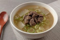 a white bowl filled with soup and meat on top of a table next to a wooden spoon