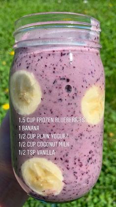 a person holding up a jar filled with blueberry and banana smoothie on top of green grass