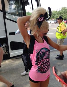 a woman in pink shirt and black shorts standing next to a bus with her hand on her head