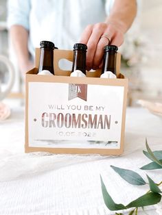 a man holding two bottles of groomsman beer in front of a sign that says will you be my groomsman?