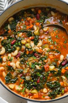 a pot filled with soup and vegetables on top of a white table cloth next to a spoon