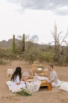 a man and woman sitting at a picnic table in the desert with food on it