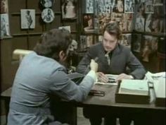 two men sitting at a desk in front of a book shelf with books on it