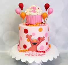 a two tiered cake decorated with pink and orange icing, topped with cupcakes and balloons