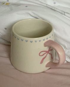 a white cup with pink handles sitting on top of a bed next to a pillow