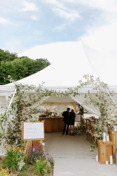 two people standing under a white tent with flowers and greenery on the side walk