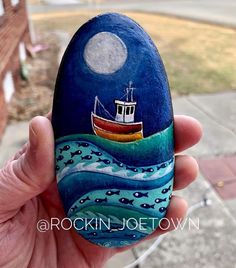 a hand holding a painted rock with a boat on the water and moon in the sky