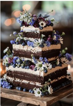 a three layer chocolate cake with blue flowers and berries on the top is sitting on a wooden platter