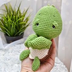 a small crocheted turtle is held in someone's hand with a potted plant behind it