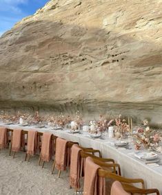 a long table is set up in front of a large rock formation with flowers and plates on it