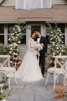 a bride and groom standing in front of a house with white flowers on the lawn