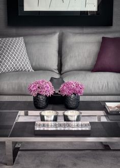 a living room with grey couches and purple flowers in vases on the coffee table