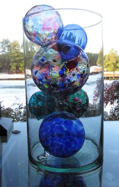 several glass balls are stacked in a vase on a table near some bushes and water