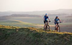 two bicyclists riding on the top of a grassy hill with rolling hills in the background