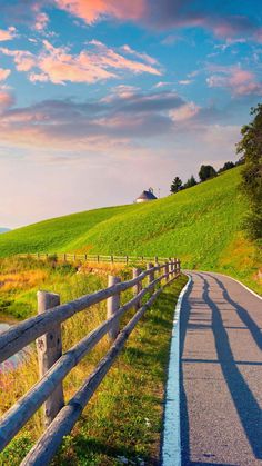 an empty road in the middle of a green field with a wooden fence around it