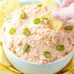 a hand dipping a chip into a bowl of dip with green onions and jalapenos