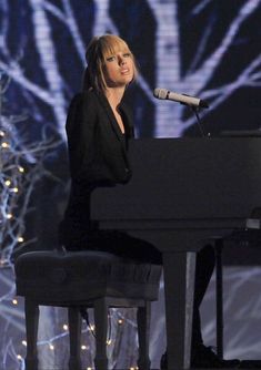 a woman sitting at a black piano in front of a microphone and christmas lights behind her