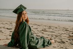 a woman in a green graduation gown sitting on the beach with her arms around her head