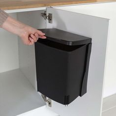 a hand is holding the door to a black trash can in a white cabinet with shelves