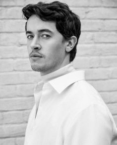 a black and white photo of a man in front of a brick wall looking at the camera