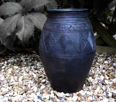 a large black vase sitting on top of a gravel covered ground next to plants and rocks