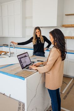 two women standing at a kitchen counter with a laptop computer on the counter and another woman in the background