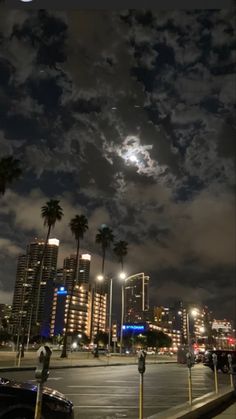 the night sky is filled with clouds and palm trees, as seen from an empty parking lot