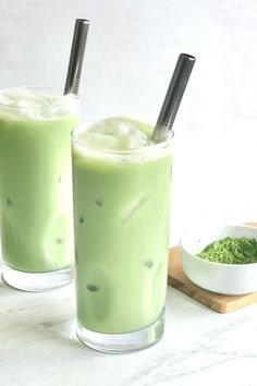 two glasses filled with green liquid on top of a white counter next to a bowl and spoon