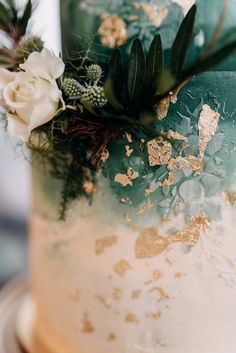 a green and gold wedding cake with white flowers on the top, surrounded by greenery