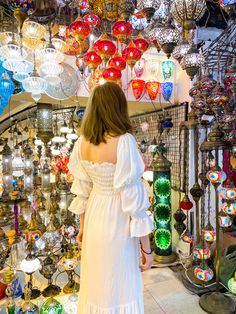 a woman in a white dress is looking at many colorful glass lamps and lights hanging from the ceiling