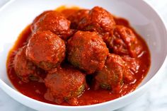 a white bowl filled with meatballs covered in marinara sauce on top of a marble counter