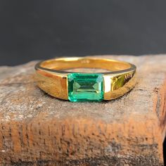 a gold ring with an emerald stone on top of a wooden block, sitting on a piece of wood