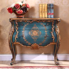 a blue and gold painted chest of drawers with flowers on top, next to bookshelves