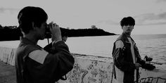 two men standing next to each other near the ocean taking pictures with their cell phones