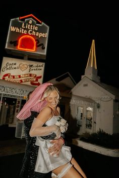 two women are hugging in front of the little white chapel sign and lit up building